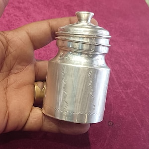 May include: A silver jar with a decorative lid and a geometric pattern etched into the body. The jar is made of metal and has a shiny finish.