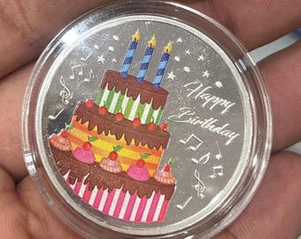 May include: A silver coin featuring a colorful birthday cake with three lit candles. The cake has layers of chocolate frosting, pink and yellow stripes, and cherries. The words "Happy Birthday" are written on the coin, along with musical notes.