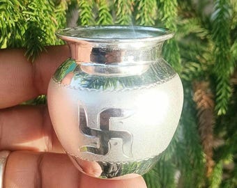 May include: A small, polished silver-colored metal pot with a wide rim and a rounded body. The exterior features a matte finish with a prominent, smooth, embossed swastika symbol centered on the front, framed by decorative horizontal bands.