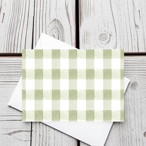 Puede incluir: Una tarjeta de felicitación con un patrón de cuadros verdes y blancos, colocada sobre un sobre blanco. La tarjeta y el sobre están sobre una superficie de madera desgastada pintada de blanco. El diseño de la tarjeta presenta un patrón de cuadros estilo acuarela.