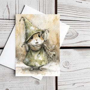 May include: A whimsical greeting card featuring a watercolor illustration of a mouse wearing a green cloak and hat, shielding itself from the rain. The mouse has a scarf and is standing in a snowy scene. The card is on a white envelope.
