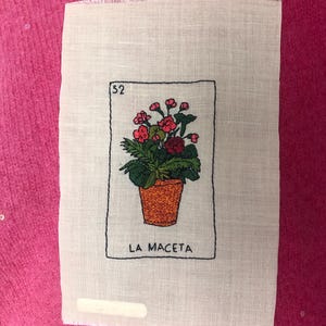 Peut inclure: Un morceau de tissu rectangulaire blanc cassé avec une broderie représentant une plante en pot aux fleurs roses et aux feuilles vertes. Le motif est encadré par un rectangle noir avec le numéro 52 et le texte LA MACETA.