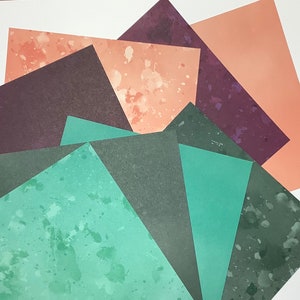 May include: An assortment of colorful paper sheets arranged in a fan shape. The papers feature various shades of green, purple, and peach, with some displaying speckled patterns. The papers are likely for crafting or scrapbooking.