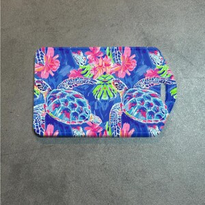 May include: Rectangular cutting board with a blue background featuring two sea turtles and pink and white tropical flowers. The board has a handle on the right side.