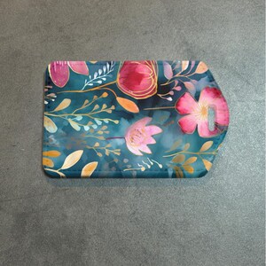 May include: A rectangular teal cutting board with a floral design. The design features pink and red flowers with gold outlines, and orange and gold leaves. The cutting board has a handle with a hole.