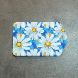 May include: A rectangular card holder featuring a daisy floral pattern. The design showcases white daisies with yellow centers against a vibrant blue background. The card holder has rounded corners and a small cutout.