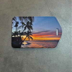 May include: A rectangular luggage tag featuring a tropical beach scene at sunset. The image shows palm trees silhouetted against a vibrant sky of orange, yellow, and blue hues. The tag has a black border and a strap hole.