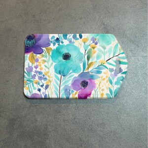 May include: A rectangular floral design with rounded edges. The design features watercolor-style flowers in shades of teal, purple, yellow, and green on a white background. The item is likely a coaster or small tray.