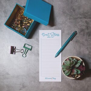 May include: A white notepad with the text "Don't Stress Make a List" in blue cursive writing. The notepad has a blue line border and a list of checkboxes. The notepad is on a gray surface with a blue pen, a green plant in a white pot, and a blue box with colorful paper clips.