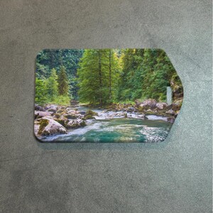 May include: Rectangular luggage tag featuring a scenic river landscape with lush green trees and rocky banks. The water is a vibrant turquoise color, and the tag has a rounded corner and a hole for attachment.