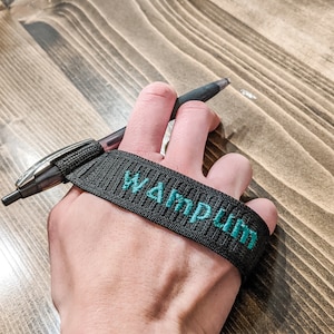 May include: A black pen with a black strap that has the word "Wampum" embroidered in teal. The pen is held in a hand, resting on a wooden surface.