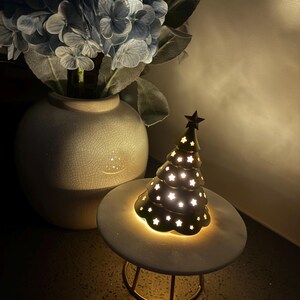 May include: A gold metal Christmas tree with a star on top and a white base. The tree is lit up with white lights and has a cutout design of stars. The tree is sitting on a white marble pedestal with a gold metal base.