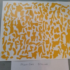 Puede incluir: Una hoja de papel blanca con un patr&oacute;n de camuflaje amarillo. El patr&oacute;n consiste en formas irregulares de varios tama&ntilde;os. El texto "Multi Cam Styling" est&aacute; escrito en la parte inferior del papel.