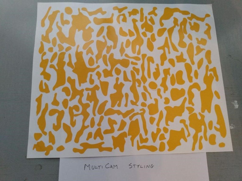 Puede incluir: Una hoja de papel blanca con un patr&oacute;n de camuflaje amarillo. El patr&oacute;n consiste en formas irregulares de varios tama&ntilde;os. El texto "Multi Cam Styling" est&aacute; escrito en la parte inferior del papel.
