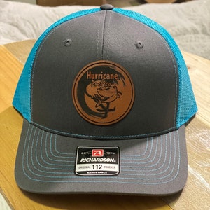 Puede incluir: Un gorro de camionero gris y turquesa con un parche de cuero marrón que dice "Hurricane Anchors" con un gráfico de dos peces. El gorro es ajustable y tiene un logotipo de Richardson.