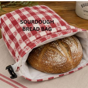 May include: A red and white checkered sourdough bread bag with the words "SOURDOUGH BREAD BAG" printed on the front. The bag is open, revealing a loaf of bread. The bag has a white interior and a black plastic clip.