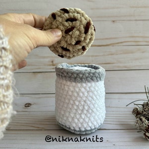 May include: A crocheted chocolate chip cookie being placed into a crocheted milk carton. The cookie is brown and tan with brown chocolate chips. The milk carton is white with a gray rim. The background is a light wood surface. The text "@niknaknits" is visible at the bottom of the image.