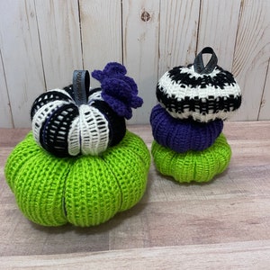 May include: Two crocheted pumpkins, one green and one stacked with black and white, purple, and green. The larger pumpkin has a purple flower on top and a tag that says "Made with Love".