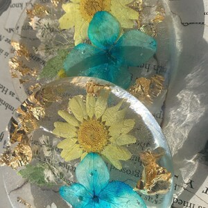 Puede incluir: Dos posavasos redondos de resina con flores prensadas y pan de oro. Los posavasos presentan una margarita amarilla y una hortensia azul.