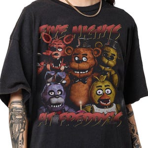 May include: Black t-shirt featuring a graphic design from the video game "Five Nights at Freddy's." The design includes the game's title in red text and several character illustrations, including Freddy, Bonnie, Chica, and Foxy.