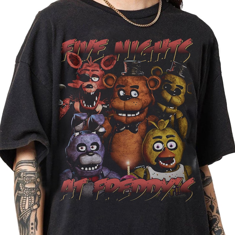 May include: Black t-shirt featuring a graphic design from the video game "Five Nights at Freddy's." The design includes the game's title in red text and several character illustrations, including Freddy, Bonnie, Chica, and Foxy.