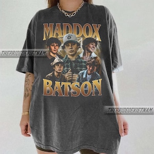 May include: Dark gray t-shirt featuring a graphic of five young men with the names "MADDOX" and "BATSON" in gold lettering. The shirt has a vintage, worn look, with the text "THEPRIDEOFVIETNAM" printed on the side.