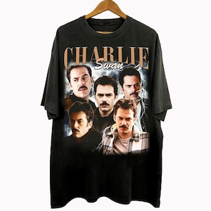 May include: Black t-shirt with a vintage-style graphic of Charlie Swan, featuring multiple images of the character. The text "CHARLIE Swan" is printed in gold across the top. The shirt is displayed on a wooden hanger.