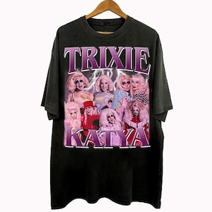May include: Black t-shirt featuring a group of drag queens with the text "Trixie Katya" in a purple retro font. The design includes a collage of images of the queens with lightning bolt graphics.