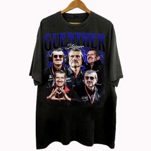 May include: Black t-shirt featuring a graphic of multiple men with the text "GUENTHER Steiner" in blue. The design has a vintage, distressed look. The shirt is hanging on a wooden hanger.