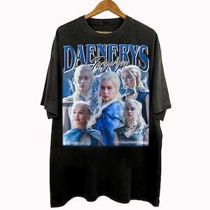 May include: Black t-shirt featuring a graphic of Daenerys Targaryen with the text "DAENERYS Targaryen" in blue. The design includes multiple images of the character with lightning bolt accents. The shirt is hanging on a wooden hanger.