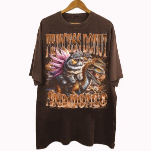 May include: Brown t-shirt with the text "PRINCESS DONUT AND MONGO". The graphic features a cat with sunglasses and a crown riding a dinosaur, with pink and purple accents.