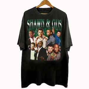 May include: Black t-shirt featuring a graphic of the cast of "Shawn & Gus" with the show's title in green. The design includes a group of men in various outfits against a green lightning background. The shirt is hanging on a wooden hanger.