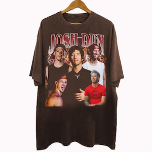 May include: Brown t-shirt featuring a vintage-style graphic of Josh Dun with the text "JOSH DUN" in red. The design includes multiple images of Josh Dun. The shirt is hanging on a wooden hanger.