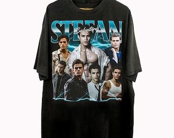 May include: Black t-shirt featuring a graphic of the name "STEFAN" in blue, with a collage of images of a man. The shirt has a vintage, distressed look, and is hanging on a wooden hanger.