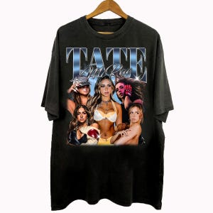 May include: Black t-shirt featuring a graphic of Tate McRae with a vintage design. The design includes the text "Tate McRae" in a stylized font, along with multiple images of the artist. The shirt is hanging on a wooden hanger.