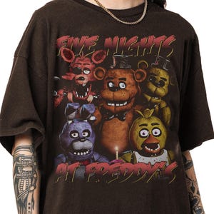 May include: Dark brown t-shirt featuring a graphic of characters from Five Nights at Freddy's. The design includes the text "FIVE NIGHTS" in red above the characters and "AT FREDDY'S" below.