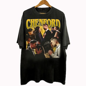 May include: Black t-shirt with a graphic featuring a group of people embracing, with the word "CHENFORD" in yellow block letters above. The design has a vintage, distressed look, and the shirt is hanging on a wooden hanger.