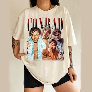May include: Cream-colored t-shirt featuring a graphic of four young men with the name "CONRAD Fisher" in a red and black font. The shirt is a casual style, likely made of cotton or a similar fabric.