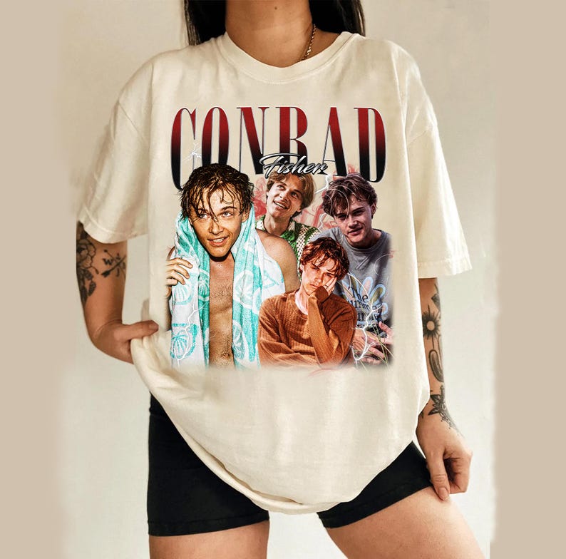 May include: Cream-colored t-shirt featuring a graphic of four young men with the name "CONRAD Fisher" in a red and black font. The shirt is a casual style, likely made of cotton or a similar fabric.