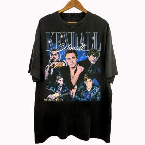 May include: Black t-shirt featuring a vintage-style graphic of Kendall Schmidt and the text "Kendall Schmidt" in a blue and white design. The graphic includes multiple images of the musician. The shirt is hanging on a wooden hanger.