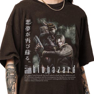 Op de afbeelding: Bruin t-shirt met een "Biohazard" graphic. Het ontwerp toont een personage met een pistool, Japanse tekst en de titel van het spel. De tekst eronder beschrijft de plot van het spel.