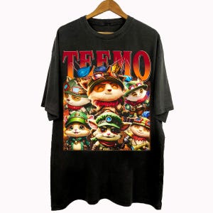 Op de afbeelding: Zwart t-shirt met een kleurrijke afbeelding van stripfiguren en het woord "TEEMO" in rood en oranje. De figuren dragen hoeden en verschillende outfits. Het t-shirt hangt aan een houten kleerhanger.