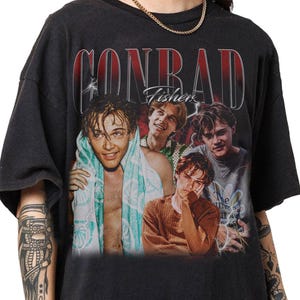 May include: Black t-shirt featuring a vintage-style graphic of four young men. The word "CONRAD" is printed in red above the images, with "Fishers" in white script below. The design has a faded, distressed look.