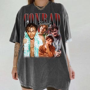 May include: Dark gray t-shirt featuring a graphic of four young men with the text "CONRAD Fisher" in a vintage style. The shirt has a relaxed fit and short sleeves. The graphic includes a photo of the young men.