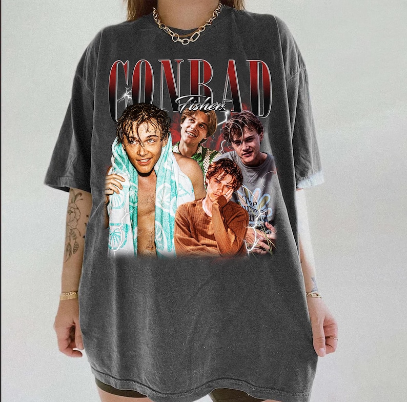 May include: Dark gray t-shirt featuring a graphic of four young men with the text "CONRAD Fisher" in a vintage style. The shirt has a relaxed fit and short sleeves. The graphic includes a photo of the young men.
