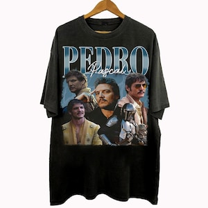 May include: Black t-shirt featuring a collage of images of Pedro Pascal with the name "PEDRO Pascal" in a blue, vintage-style font. The shirt is hanging on a wooden hanger.