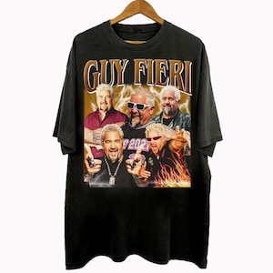 Op de afbeelding: Zwart T-shirt met een afbeelding van Guy Fieri en de tekst "GUY FIERI" in goud. De afbeelding bevat meerdere afbeeldingen van Guy Fieri. Het shirt hangt aan een houten kleerhanger.
