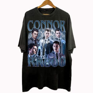Könnte beinhalten: Schwarzes T-Shirt mit einer Grafik von fünf Männern und dem Text "CONNOR" darüber und "RK800" darunter in einer blauen, verblichenen Schrift. Das Design hat einen Vintage-Look.