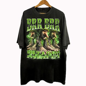 Może przedstawiać: Czarny t-shirt z graficznym wzorem przedstawiającym trzy fantastyczne stworzenia z zielonymi liśćmi i napisem "BRR BRR PATARIM" w kolorze zielonym. Koszulka wisi na drewnianym wieszaku.