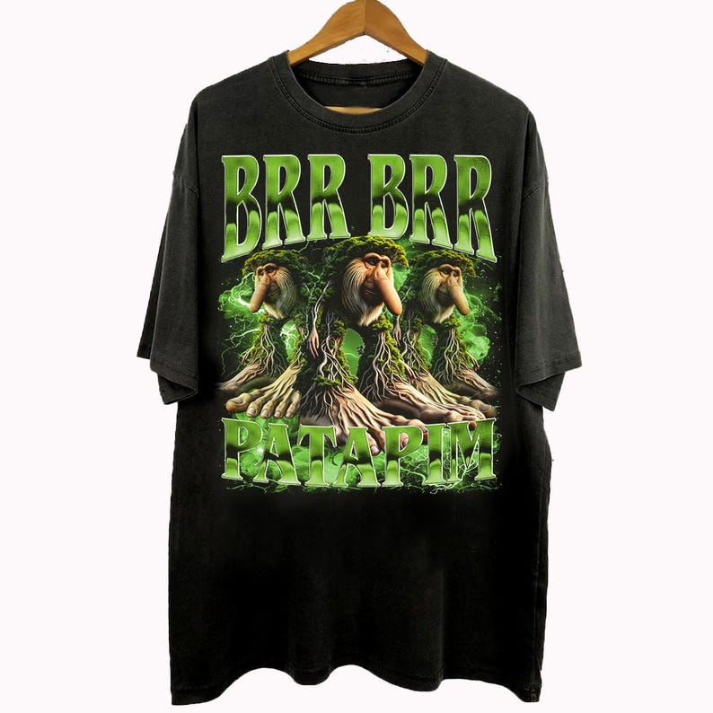 Może przedstawiać: Czarny t-shirt z graficznym wzorem przedstawiającym trzy fantastyczne stworzenia z zielonymi liśćmi i napisem "BRR BRR PATARIM" w kolorze zielonym. Koszulka wisi na drewnianym wieszaku.